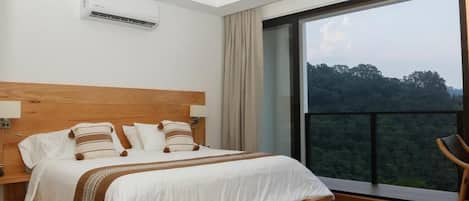 A bedroom with a large bed, a wooden headboard, a nightstand, a chair, and a view of a forest through a balcony.