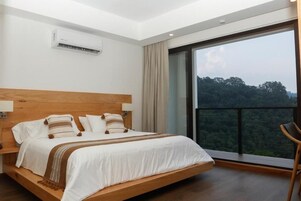 A bedroom with a large bed, a wooden headboard, a nightstand, a chair, and a view of a forest through a balcony.