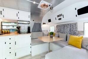 Interior d'una caravana compacta amb cuina americana, zona d'estar i una taula petita amb una planta.