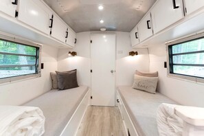 A compact, well-lit room with two beds, each with a pillow and a bedside table. There are windows on both sides, allowing natural light and views of the outdoors. - Glamping in an Airstream: Beautiful Accommodation near Tobermory, Canada (Northern Bruce Peninsula)