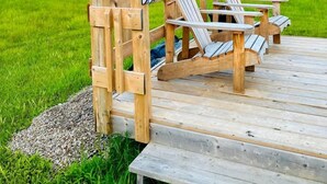 Puinen terassi, jossa on Adirondack-tyyliset puutarhatuolit ja pieni pöytä, ja jolta avautuu näkymä vehreälle niitylle ja vesistölle.