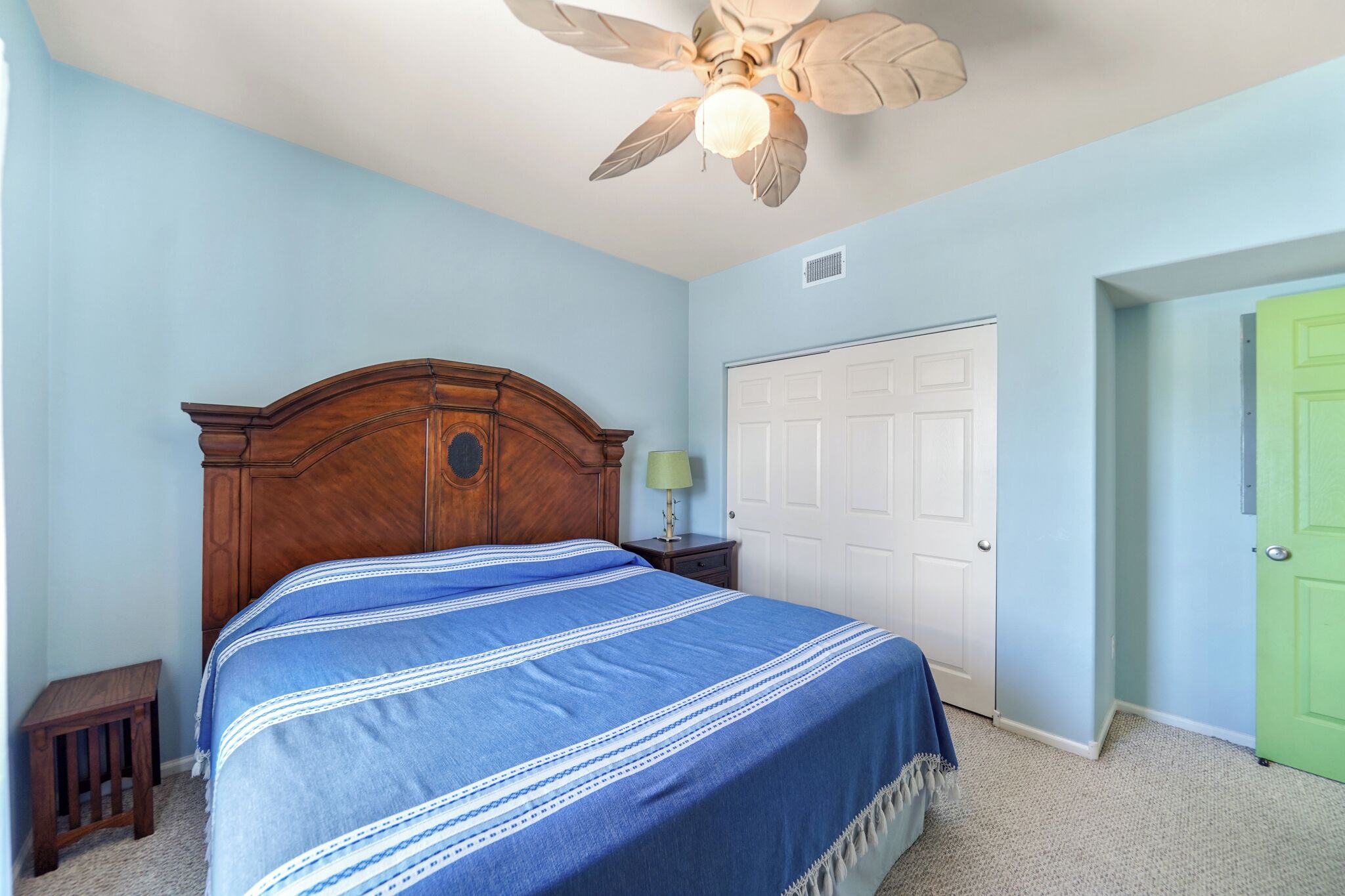 A bedroom with a bed, a nightstand, a lamp, a ceiling fan, a white door, and a green door.