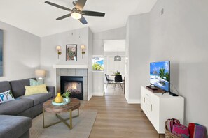 Uma sala de estar moderna com lareira, ventilador de teto, área de refeições e TV sobreposta a um armário branco.