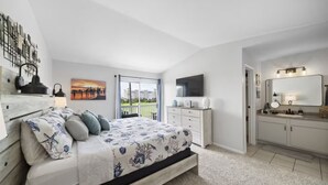 A bedroom with a bed, a dresser, a television, and a bathroom with a mirror and sink. - Palms Escape | Ocean View Condo | Pool | Pet-Friendly Retreat (Galveston)