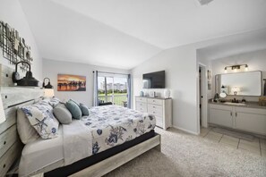 A bedroom with a bed, a dresser, a television, and a bathroom with a mirror and sink. - Palms Escape | Ocean View Condo | Pool | Pet-Friendly Retreat (Galveston)