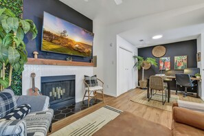A living room with a fireplace, a television mounted on the wall, and a dining area with a table and chairs. - 2 Workspaces + Pool | Couples’ Galveston Escape (Galveston)