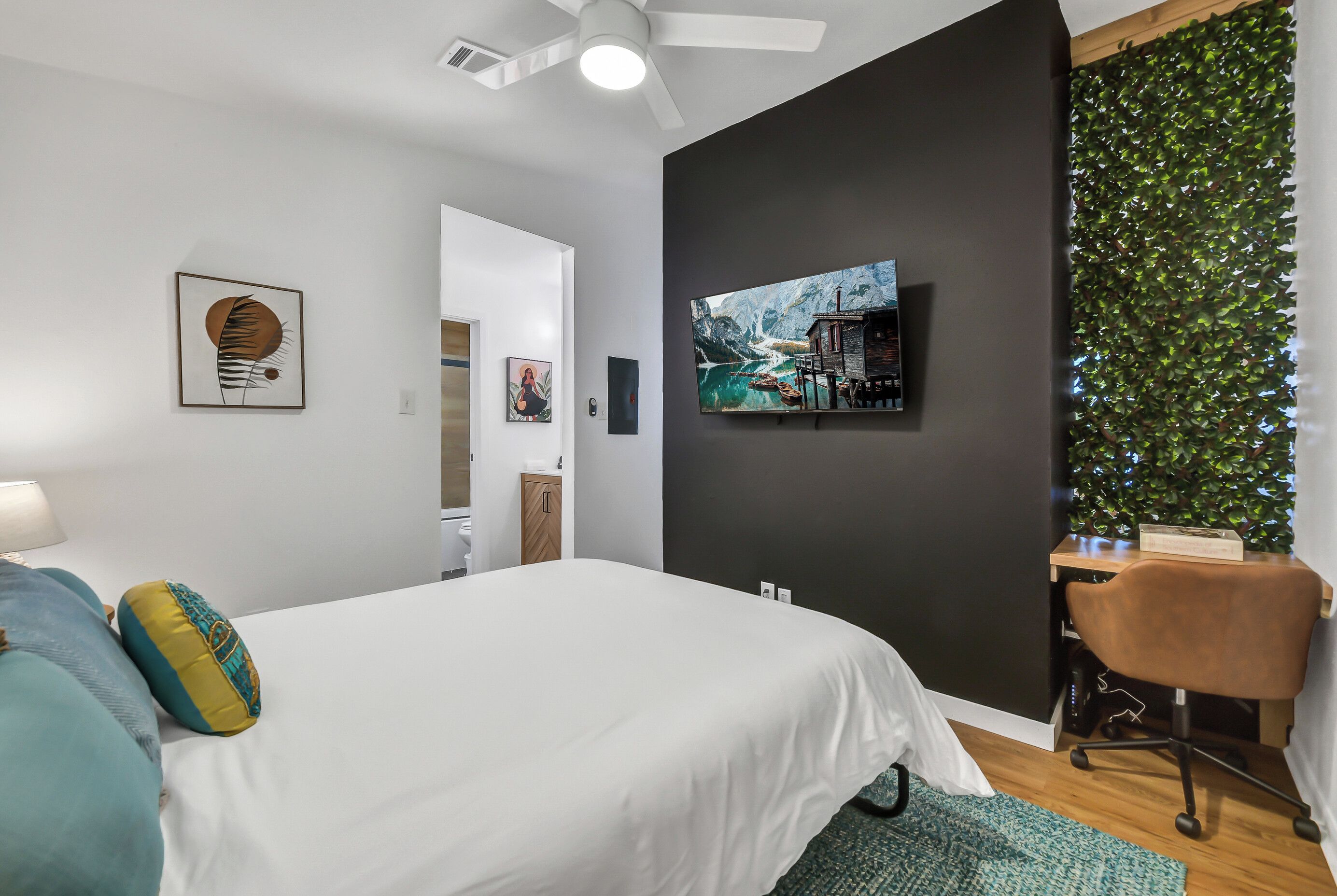 A modern bedroom with a bed, a desk, and a chair. There is a painting on the wall and a plant on the right side.