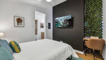 A modern bedroom with a bed, a desk, and a chair. There is a painting on the wall and a plant on the right side.