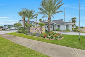 A residential area with a sign for 'The Palm at Cove View' and a well-maintained lawn with palm trees. - 2 Workspaces + Pool | Couples’ Galveston Escape (Galveston)