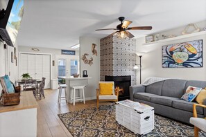 Uma sala de estar com lareira, um sofá cinzento, um tapete estampado e um ventilador de teto.