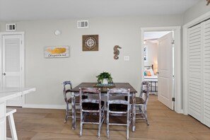 A dining area with a wooden table and chairs, a wall-mounted compass, and a framed quote. - The Salty Siren | Mermaid-Themed Condo | Pool Access | Private Patio (Galveston)