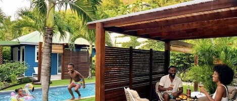 A pool area with a wooden pergola, lounge chairs, and a dining table with guests.