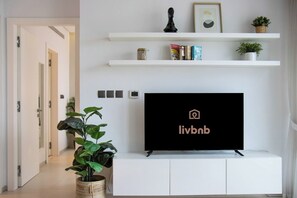 A modern living room with a flat-screen TV on a white console, a potted plant, and white shelving with decorative items. - 1B Walk to Metro, Marina Mall with Skyline Views (Dubai)