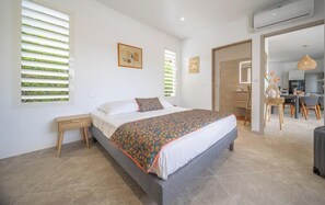 A bedroom with a large bed, a bedside table, a window with blinds, and a view into the adjacent kitchen area.