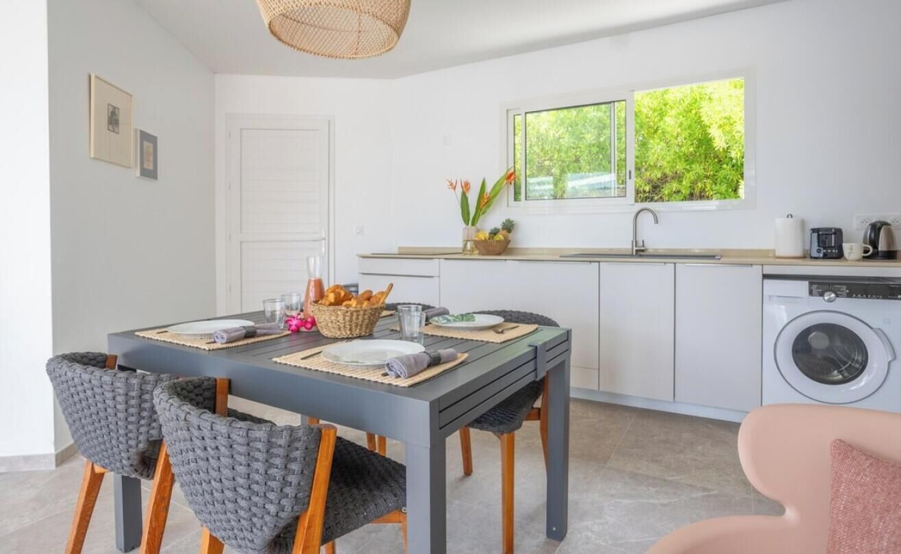 A modern kitchen with a dining area, a washing machine, and a window with a view of greenery.