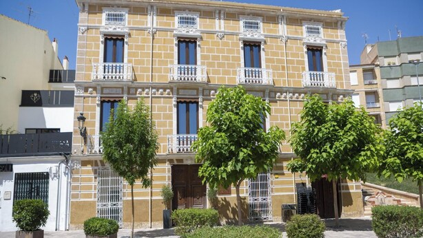 Un edificio de varios pisos con balcones, entrada protegida por una verja y zonas verdes circundantes.
