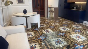 Una sala de estar moderna con una mesa redonda y sillas blancas, una zona de cocina con encimera oscura y un suelo estampado.