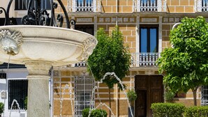 Una fuente de diseño clásico, rodeada de plantas en macetas y un edificio con balcones y ventanas.