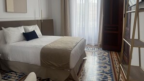 Habitación de hotel con cama, un ventanal con cortinas translúcidas, piso estampado y puerta de madera.