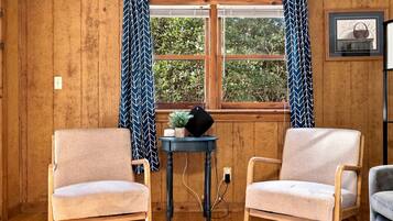 Une pièce aux murs en bois, avec deux fauteuils, une petite table sur laquelle est posée une plante, et une fenêtre avec des rideaux.
