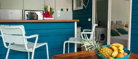 Cocina compacta con una mesa y sillas de madera, un frutero y un ventilador.