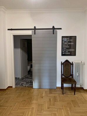 A room with a sliding door, a radiator, a chair, and a framed list of names on the wall. - Acropolis Oasis | Designer Home by the Acropolis (Athina)
