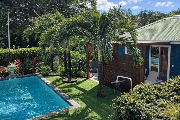 A swimming pool surrounded by lush greenery, a wooden deck, and a small building with a green roof.