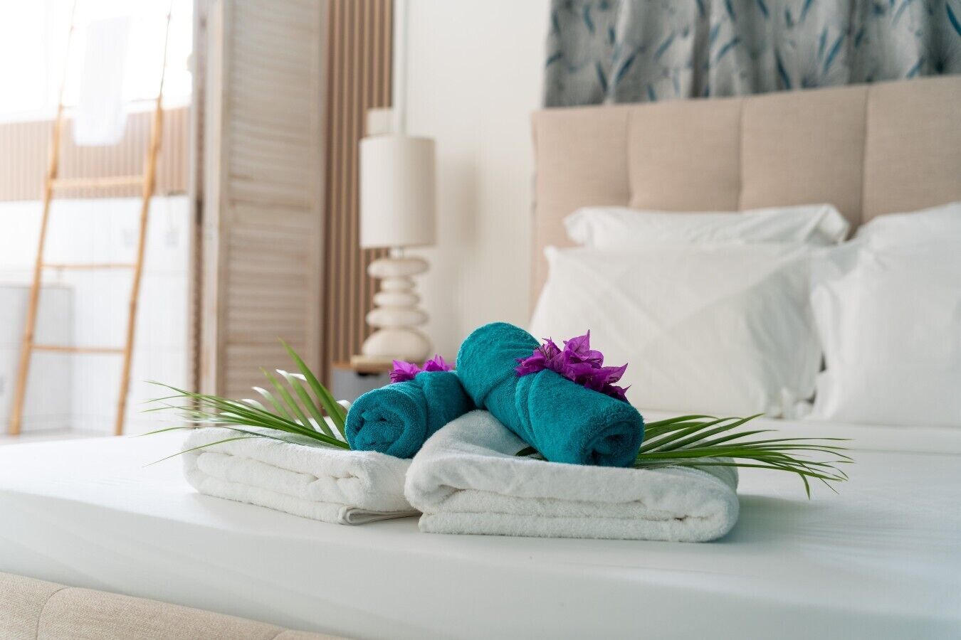 Una cama bien hecha con toallas enrolladas y una flor decorativa, una mesita de noche con una lámpara, y una ventana con cortinas.