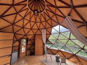 A geodesic dome room with a wooden floor, a small table, chairs, a refrigerator, and a large window offering a view of the landscape.