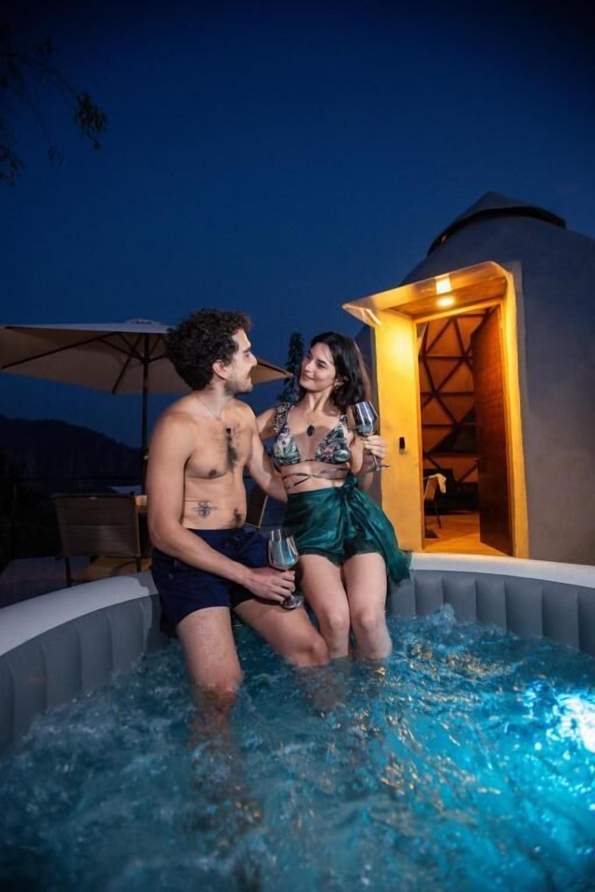 Una pareja en un jacuzzi con vistas al cielo nocturno y una edificación al fondo.