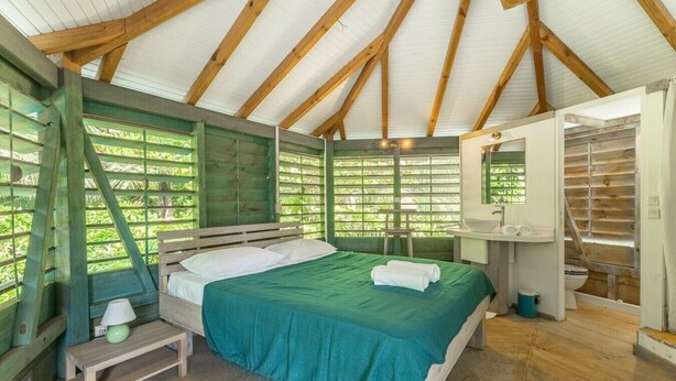A room with a bed, a small table, a bench, a window with a view of greenery, and a bathroom with a sink and toilet. - Domaine De L'Anse Noire, right on the water (Les Anses-d`Arlet)