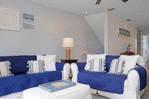 A modern living room with a blue and white sofa set, a side table with a lamp, and a wall-mounted sign. - Barrier Dunes 542 - 55 - Relax N' Moore (Port St. Joe)