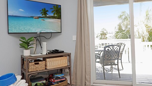 Quarto com televisão exibindo uma cena de praia tropical, uma pequena planta, um vaso branco e uma estante com diversos objetos, incluindo uma caixa rotulada 'Rummikub Six Player'.