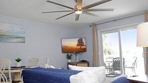 Quarto de hotel com cama, escrivaninha, cadeira, ventilador de teto, janela com cortinas e um quadro na parede.
