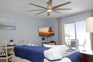 Ein Hotelzimmer mit Bett, Schreibtisch, Stuhl, Deckenventilator, einem Vorhangfenster und einem Bild an der Wand.