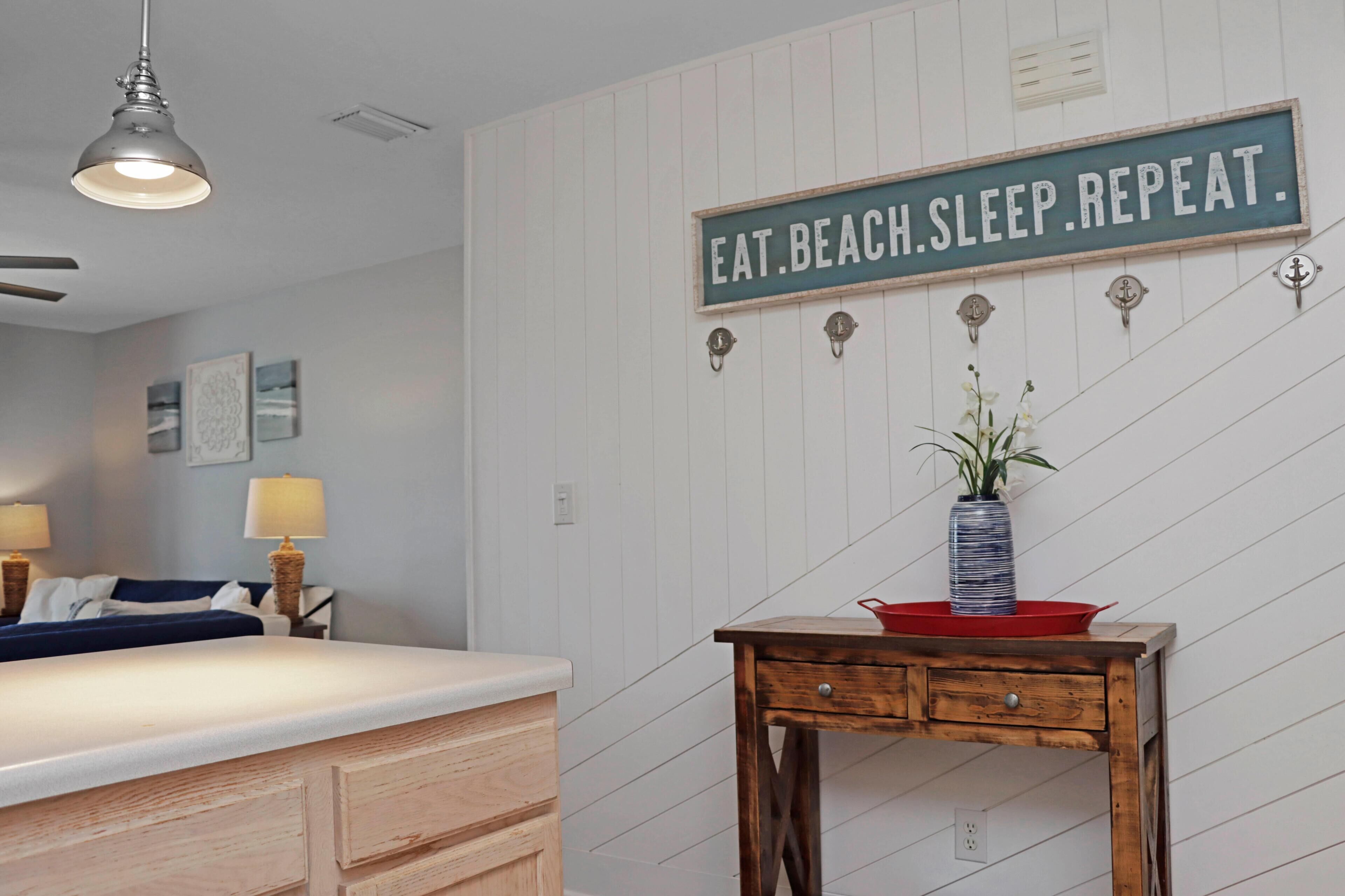 A kitchen with a wooden table, a sign on the wall, and a bed in the background.