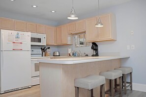 A modern kitchen with wooden cabinets, a white refrigerator, a microwave, a toaster, and a coffee maker. There are three bar stools and pendant lights above the island. - Barrier Dunes 542 - 55 - Relax N' Moore (Port St. Joe)