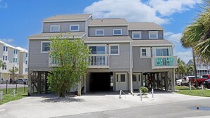 Um edifício residencial de vários andares com entrada frontal, uma pequena árvore e área de estacionamento.