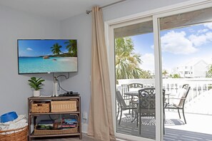 A room with a television displaying a beach scene, a wicker shelf with storage baskets, and a view of a balcony with outdoor furniture. - Barrier Dunes 542 - 55 - Relax N' Moore (Port St. Joe)