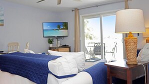 Quarto de hotel com cama, ventoinha no teto, televisão, janela com vista para palmeiras e mesa com abajur.
