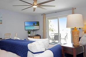 Ein Hotelzimmer mit einem Bett, einem Deckenventilator, einem Fernseher, einem Fenster mit Blick auf Palmen und einem Tisch mit einer Lampe.