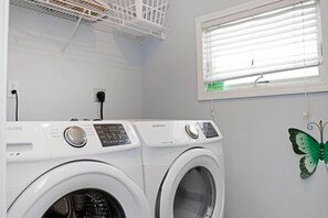 A laundry room with two front-loading washing machines, a ceiling vent, a window with blinds, and a green butterfly decoration on the wall. - Barrier Dunes 542 - 55 - Relax N' Moore (Port St. Joe)