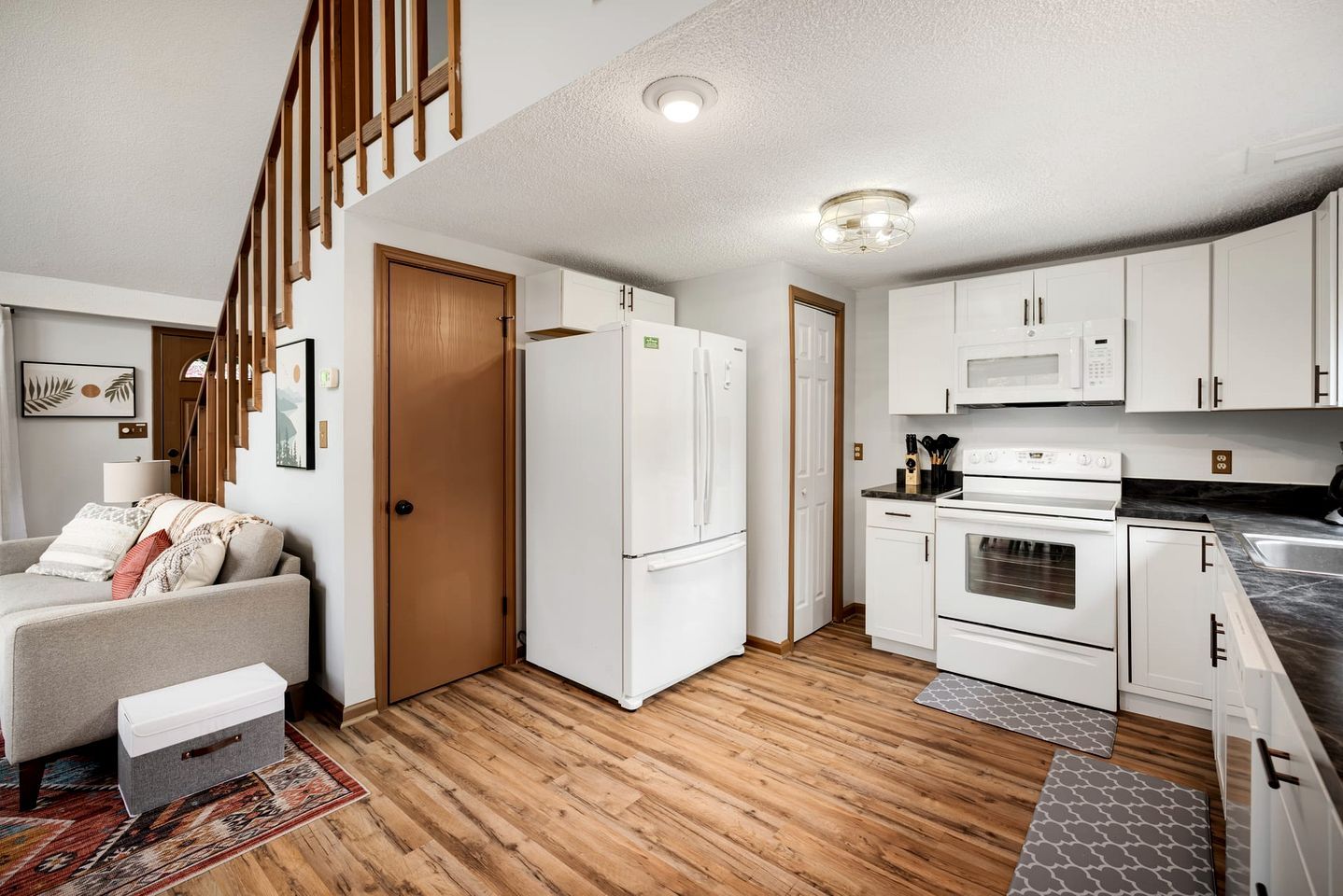 A compact kitchen with white appliances, a wooden floor, and a staircase leading to another level.