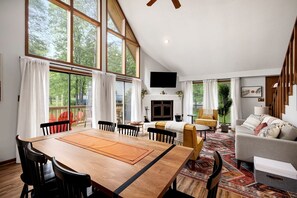A spacious living area with a large wooden dining table, comfortable seating, a fireplace, and large windows offering a view of the outdoors.
