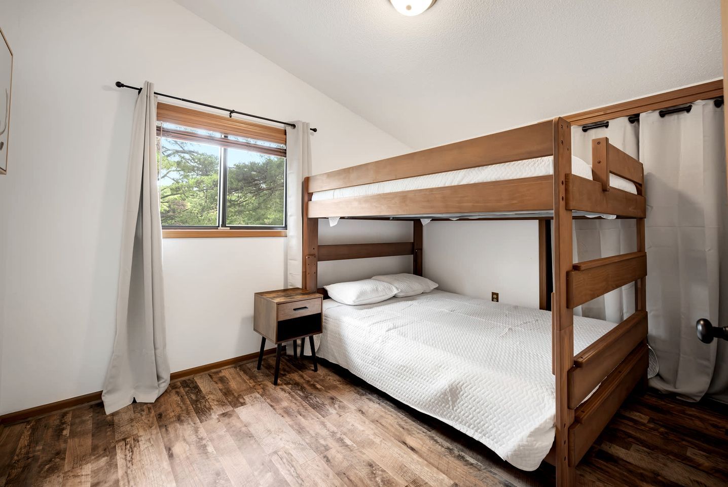 A bunk bed room with a wooden headboard, a bedside table, and a window with curtains.