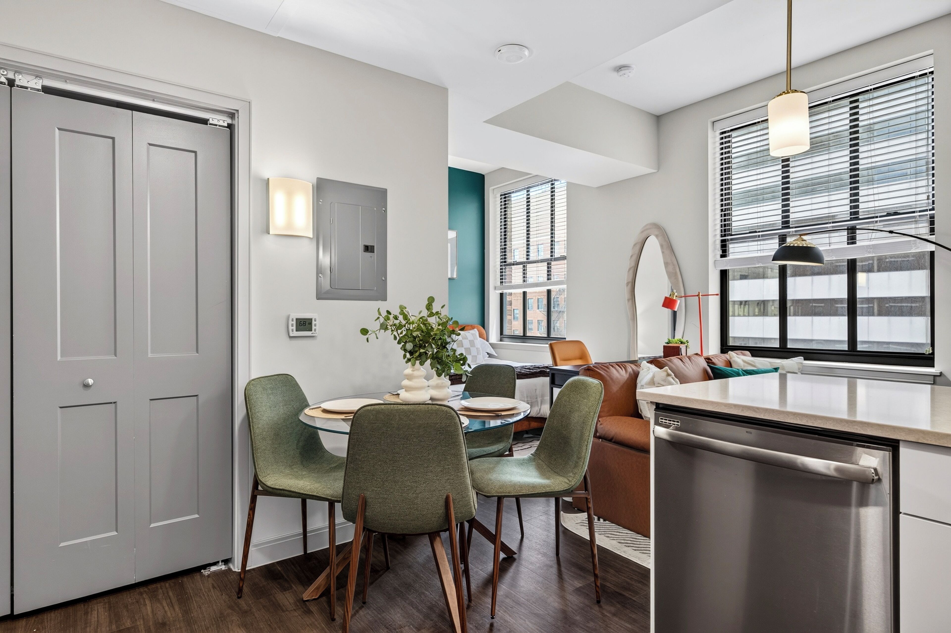 A modern kitchen with a dishwasher, a dining area with a round table and chairs, and a view of the city through large windows.