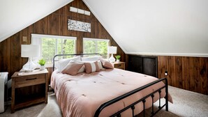 Un dormitorio con techo inclinado, paredes de paneles de madera, una cama con colcha rosa, dos mesitas de noche con lámparas y una ventana con vistas a árboles.