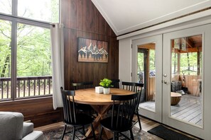 A dining area with a wooden table and chairs, a painting on the wall, and a view of the outdoors.