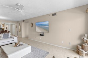 Ein geräumiges Wohnzimmer mit Deckenventilator, einem großen Flachbildfernseher an der Wand, einem weißen Sofa und einem Couchtisch.