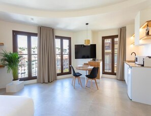Ein modernes Hotelzimmer mit Essbereich, Flachbildfernseher, einer kleinen Küche und großen Fenstern mit Vorhängen.